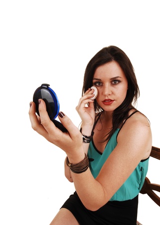 A young brunette woman sitting and applying makeup to her face, holdingup the mirror and looking into the camera for white background の写真素材