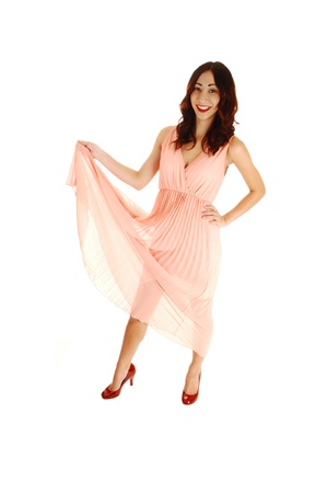 A happy young woman in a light pink dress, lifting the dress up, with redhigh heels and long brunette hair, standing for white background の写真素材