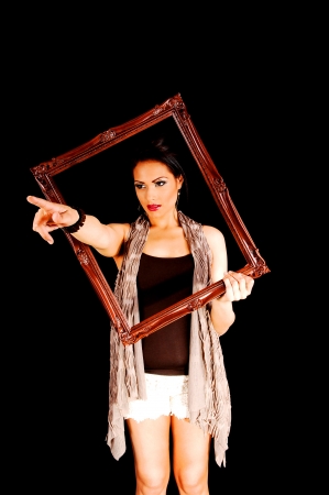 A lovely young woman in white shorts, brown t-shirt and a scarf, holding abrown picture frame for black background, pointing with her finger の写真素材