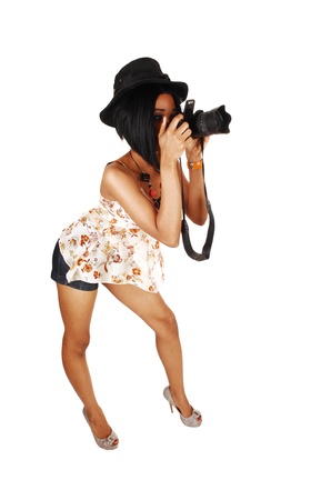 A slim young woman standing for white background in shorts and heelswith a hat on her head taking pictures with her camera の写真素材