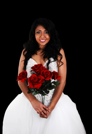 A happy young Hispanic woman in a white wedding dress standing forblack background, holding a bunch of red roses in her hand の写真素材