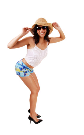 A lovely young and slim woman in shorts and white top holding a straw hat on her head, with sunglasses, isolated on white background の写真素材