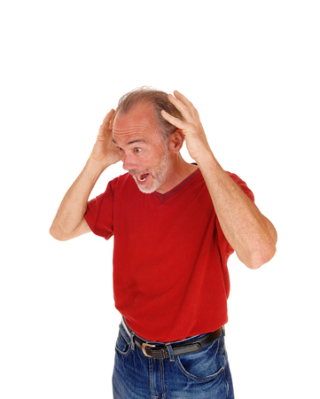 A older man in a red t-shirt and jeans standing with his hands on his head
screaming, isolated for white background.の写真素材
