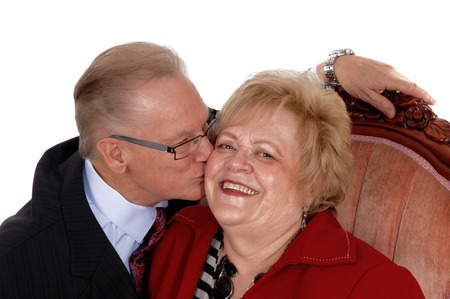 A closeup image of a seventies year old couple, the wife sitting in a pinkarmchair and her husband...