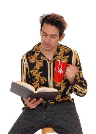 A tall young man sitting on a chair in the studio, reading his book and
holding a mug with coffee, isolated for white backgroundの写真素材