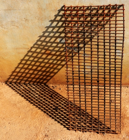 Old rusty grilling grate leans against a wall with shadows on the ground and on the wall in the shape of small rectangles の写真素材