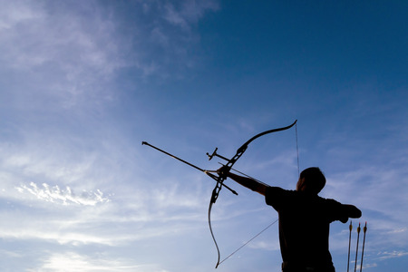 A silhouette of an archer drawing his bow and aiming upwards with deep blue sky and white clouds as backgroundの写真素材