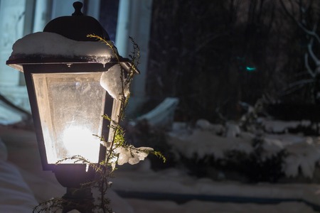 Lantern with snow and a branch of a Christmas tree glowing yellow light. Snow on the lantern, winter evening in the park.の写真素材