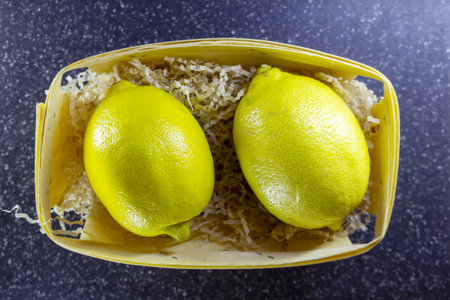 Two bright yellow lemons arranged in a rustic wooden basket with shredded paper filling