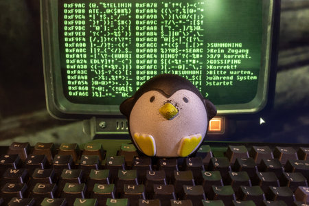 A cute toy penguin sits on a computer keyboard with green matrix code on the screenの写真素材