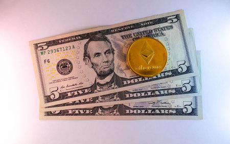 A golden ethereum coin resting on a stack of american five dollar billsのeditorial素材