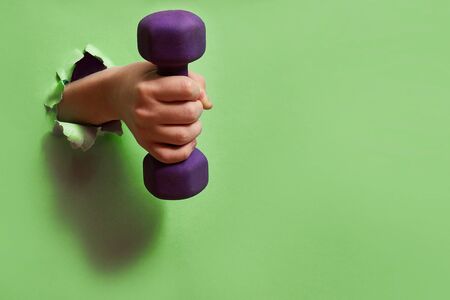 Dumbbell in hand through a hole on green paper. The concept of an active lifestyle, health and sport. Minimalism, copy space.の写真素材