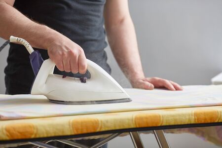 Men's hands hold a white iron and iron on the ironing board. Close-up. Faceless. Copy space. Domestic duties are performed by men.の写真素材
