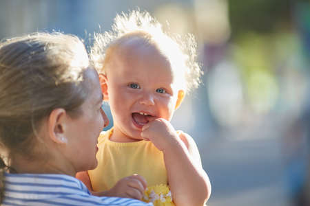 Happy smiling baby in mothers arms. Beautiful sunbeams on the street on a summer day.の写真素材