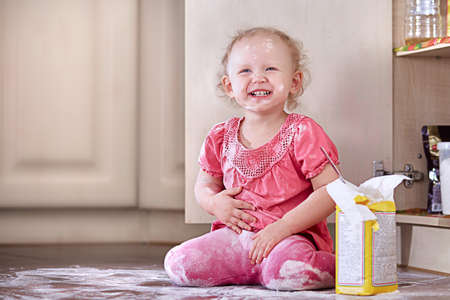Playful laughing little baby girl smeared in flour sits on the kitchen floor.の写真素材
