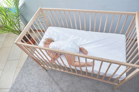 A baby sleeps on its stomach in its wooden cradle. spacious, bright room. Date palm tree under window carpetの写真素材