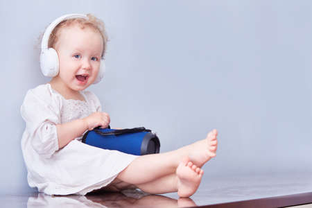 Little cute girl with a music speaker in headphones on her curly head. White dress and light back.の写真素材