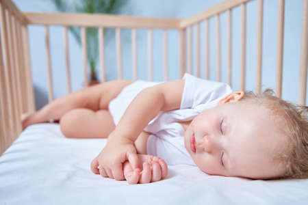 A cute baby in white clothes sleeps in a wooden crib in a bright, spacious room.の写真素材