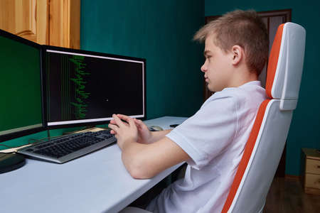 A computer programmer student sits at a computer in his room at a desk on a computer chair.の写真素材