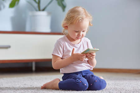 child girl look smart phone and sits on the gray carpet in living room. Child development Concept.の写真素材