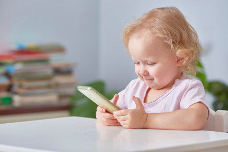 child girl is sitting at a table with a phone in her hands and smiling while looking at a smartphone.の写真素材
