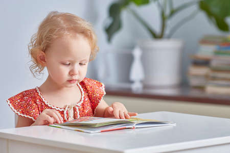 small child is reading a large book sitting at a table in a bright room.の写真素材
