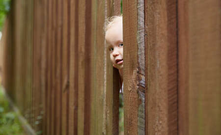 child looks out through a hole in a brown wooden fenceの写真素材