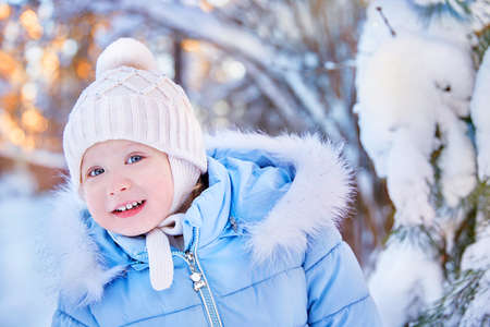 portrait of a smiling child looking into the frame into the camera. Winter landscape with a pine tree in the snow. The baby is dressed in a winter jacket with fur and a warm knitted hatの写真素材