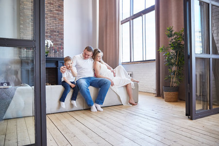 happy family with a small child and a pregnant wife is sitting on the sofa in the living room.の写真素材