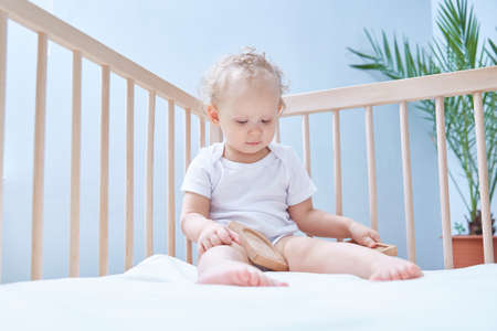 one-year-old baby is sitting in his crib in white clothes in a white room and playing a wooden pyramidの写真素材