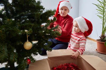 Merry Christmas and Happy Holidays. Grandmother and granddaughter decorate a Christmas tree at home. lifestyleの写真素材