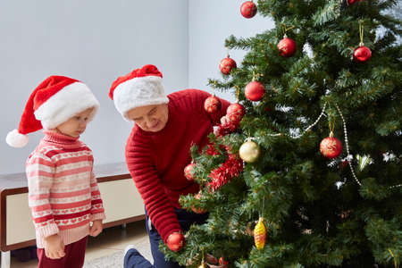 grandmother and granddaughter in Santa hats and red sweaters dress up a Christmas tree with Christmas toysの写真素材