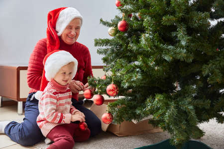 cheerful grandmother and her cute granddaughter girl decorate the Christmas tree. A grandmother and a small child are having fun near a tree indoors.の写真素材