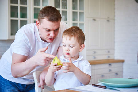 Dad and his little son are discussing an apple and healthy food in a spacious, bright kitchenの写真素材