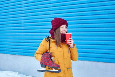 smiling young woman with skates over her shoulder and a paper cup of coffee near the bright blue wall of the ice arena stadiumの写真素材