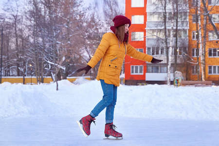 young woman on ice skates in a bright yellow jacket in winter against the background of colored high-rise buildingsの写真素材
