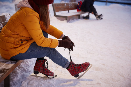 young woman in a yellow jacket ties her shoelaces on ice skates while sitting on a bench on a cold winter eveningの写真素材