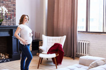 pregnant girl is standing in the living room and is touching her belly.