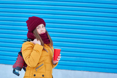 girl with a disposable cup of coffee and skates at the ready over her shoulder on a blue backgroundの写真素材