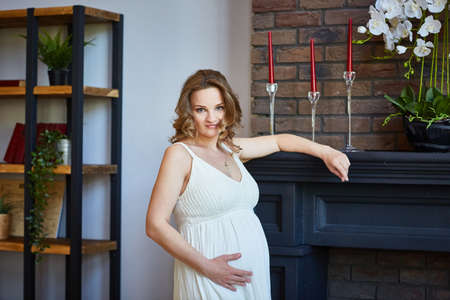 Happy pregnant young girl in the living room of her house strokes her stomach, looks at the camera and smilesの写真素材