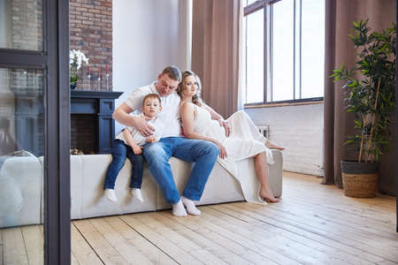Caucasian family with a small baby boy and a pregnant wife with a belly in a living room with a wooden floorの写真素材