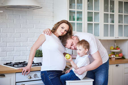 family of three with a pregnant woman and a young son are hugging in a large white kitchen.の写真素材