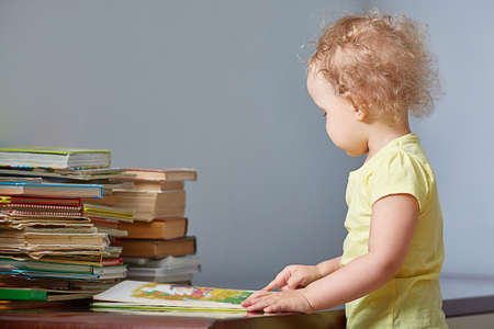 Child looks at the book. Little boy or girl reads a book and points at the book with his finger. A large stack of books next to it. Copyspace.の写真素材