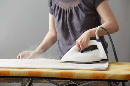 female hands are holding a white iron and iron on an ironing board. Close-up. Faceless. Copy space on gray background. Household duties and home careの写真素材