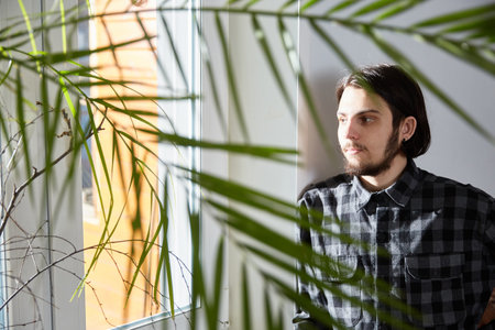 Handsome serious young bearded man standing at the window at home, looking away.の写真素材
