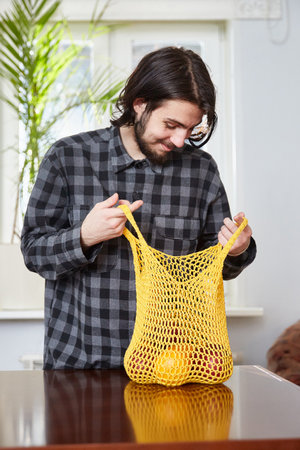 Young man holding reusable mesh bag of fresh oranges and applesの写真素材