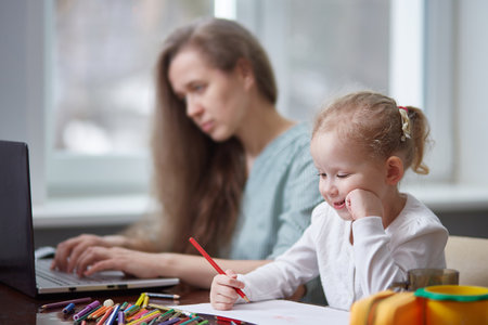 A freelance woman working at home with a child. Focus on the child. The girl draws with pencils, the mother works. Freelance, mothers Day concept.の写真素材