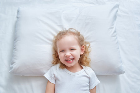 A cute curly-haired girl is lying on a white pillow smiling and looking at the camera. mock upの写真素材