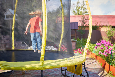 A small child jumps on a trampoline with a safety net. Happy girl outdoors in the yard of the house.on a sunny summer day.の写真素材
