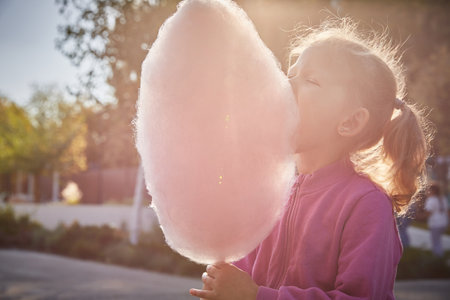 Cute curly-haired little girl eats cotton candy on a sunny summer evening in the park. Close-up.の写真素材